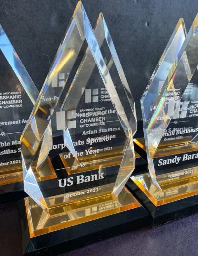 Close-up of three crystal awards with engraved text, including "US Bank" and "Sandy Barajas," on a dark background.