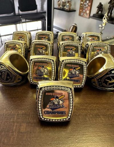 Collection of championship rings with "Pinto Division Ballers Champions" engraved, displayed on a table.