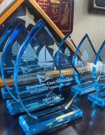 Seven blue glass sailboat trophies from the Coronado Cays Yacht Club are displayed on a shelf.